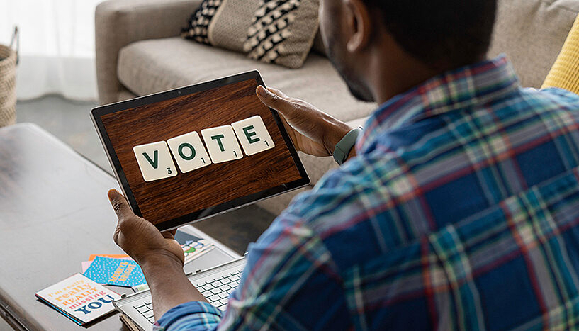 Ein Mann von hinten, am Sofa sitzend und ein Tablet in der Hand haltend. Am Bildschirm sind die Buchstaben "V-O-T-E" [engl. "wählen"] sichtbar.