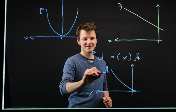 Mann zeichnet mit dafür geeigneten Stiften mathematische Formeln und Vektoren auf transparente Glastafel. Er trögt einen Mittelblauen Strickpullover - Der Hintergrund ist dunkel.