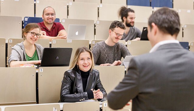Studierende sitzen im Hörsaal auf gestaffelten Sitzreihen und hören einem Vortrag zu. Einige haben Laptops vor sich, eine Frau in der vorderen Reihe lächelt dem Vortragenden zu.