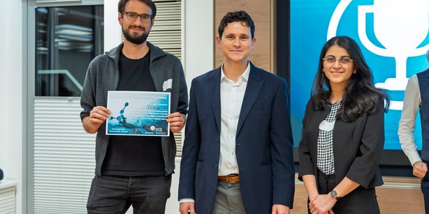 Three individuals standing indoors during a presentation event; one person holds a certificate featuring quantum-themed graphics, while a large screen in the background displays a trophy icon.