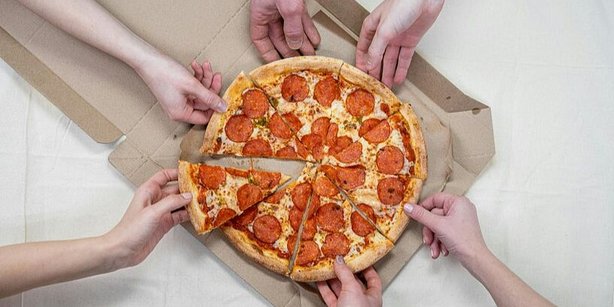 Zu sehen sind sechs Arme mit Händen, die nach einer Salami-Pizza im Zentrum des Bildes greifen.