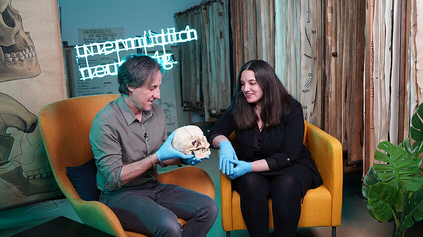Katerina Douka und Tom Higham sitting in yellwo chairs, talking to each other. Tom is holding a skull in his hands