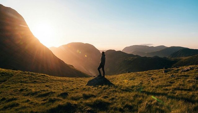 Zu sehen ist eine Person unbekannten Geschlechts auf einer Wiese mit Bergpanorama. 