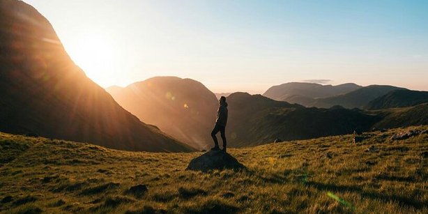 Zu sehen ist eine Person unbekannten Geschlechts auf einer Wiese mit Bergpanorama. 