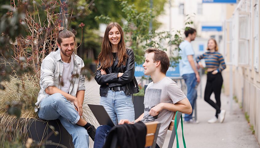 Students stand around a bench in the campus. They are talking to each other.