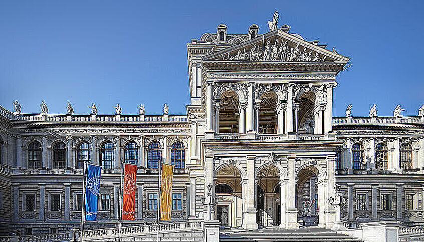 (© Alex Schuppich) Foto vom Hauptgebäude der Universität Wien. Außenansicht - Frontansicht - im Vordergrund wehen 3 bunte Fahnen.
