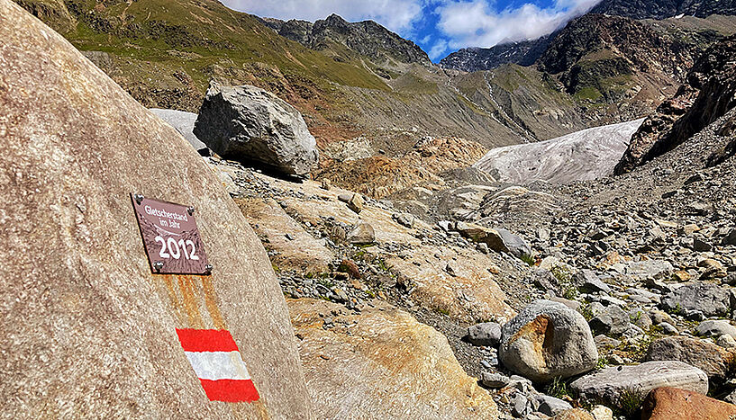Ein Schild mit der Aufschrift "Gletscherstand im Jahr 2012" prangt an einem Felsen in einer Felslandschaft in den Bergen. Weit im Hintergrund ist die Gletscherzunge sichtbar.