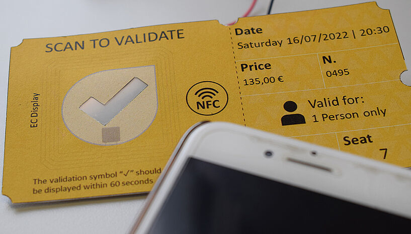 Ein gelbes Ticket mit einem hakenförmigen Ausschnitt, der beim Scannen die Farbe ändern kann. Darauf liegt ein Smartphone.