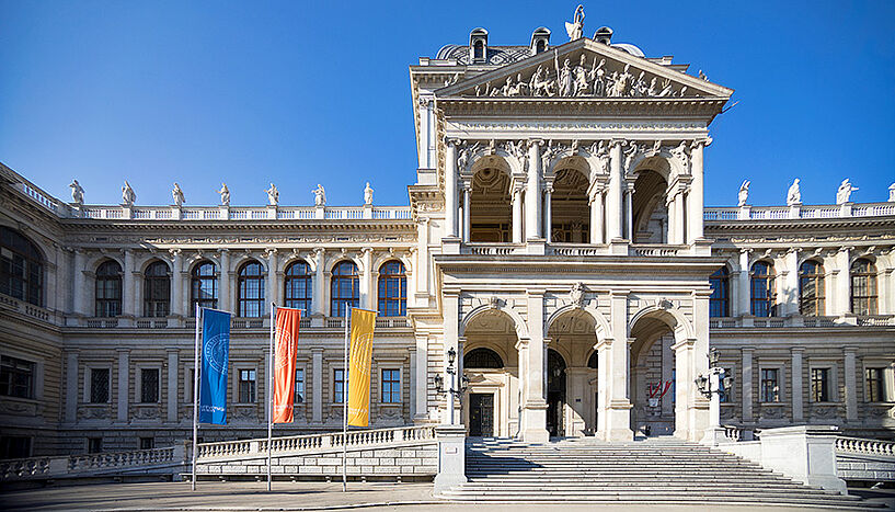 Außenansicht des Hauptgebäudes der Uni Wien im Sonnenschein und drei Flaggen die blau, geld und orange sind.