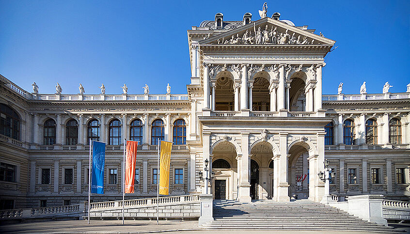 (c) Alex Schuppich Außenansicht des Hauptgebäudes der Uni Wien im Sonnenschein und drei Flaggen die blau, geld und orange sind.