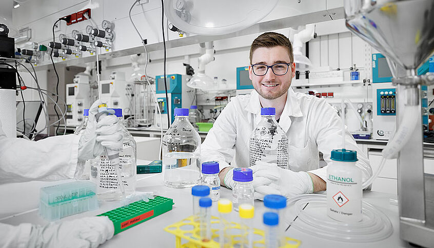Daniel im Labor mit weißen Kittel und vielen Flaschen und Gläsern rundherum