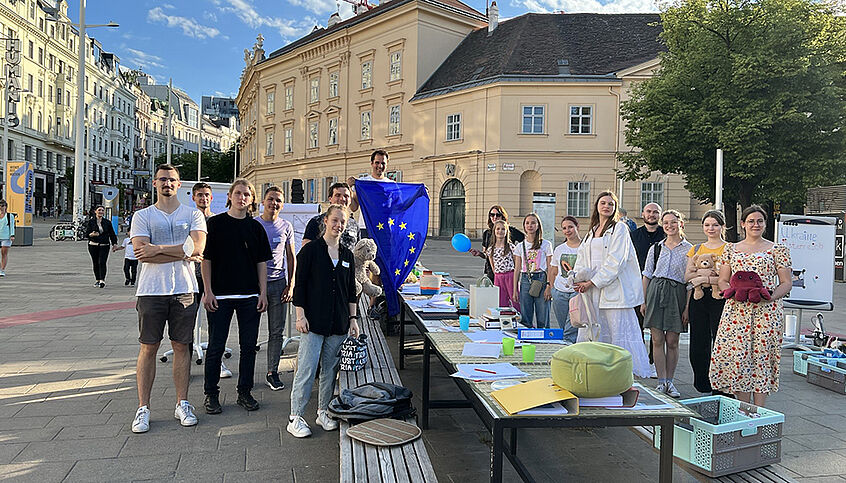© Projekt MmF Eine Gruppe junger Menschen steht um einen Tisch auf einem öffentlichen Platz (Museumsplatz, Wien). Einer hält eine EU-Flagge hoch. Auf dem Tisch zahlreiche Lernmaterialien.