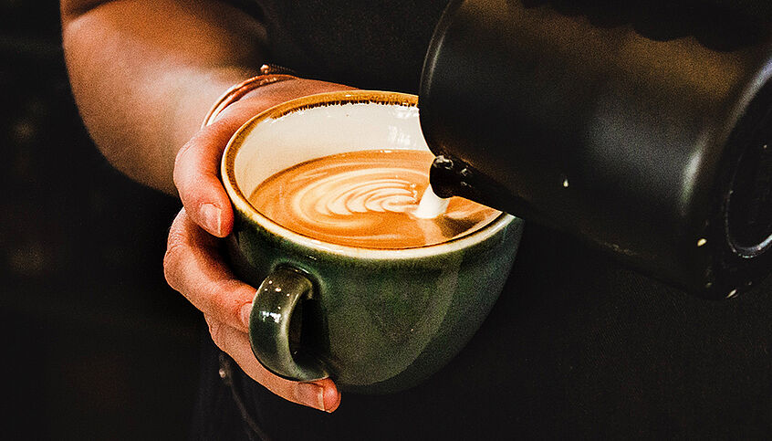 @ Louis Hansel via Unsplash Eine Hand hält ein Kaffeehäferl, in das gerade Milchschaum gegossen wird.