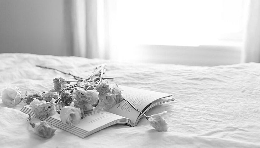 Vertrocknete Rosen liegen auf einem aufgeschlagenen Buch auf einem ordentlich gemachten Bett. Eine Schwarzweiß-Fotografie.