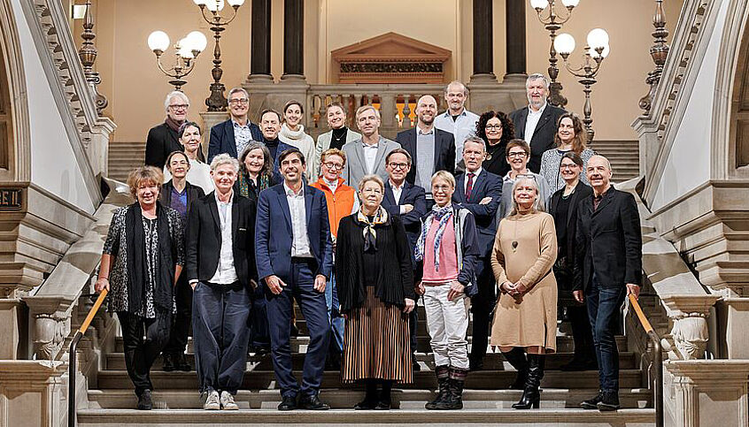 26 Personen posieren für ein Gruppenfoto auf der Prunkstiege der Universität Wien.