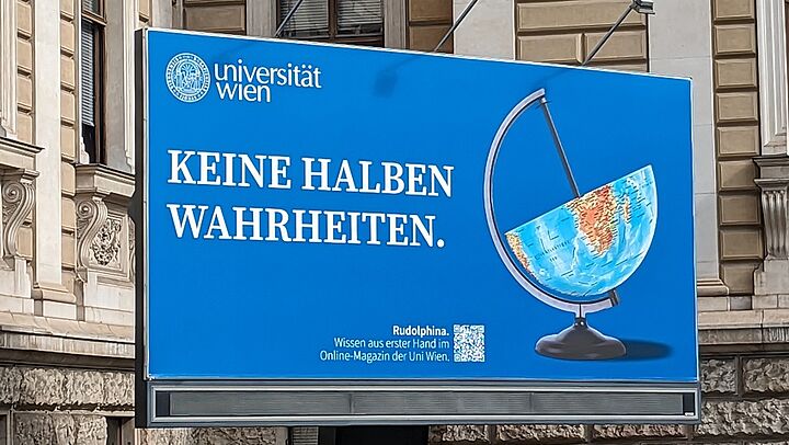 Foto vom Billboard vor dem Hauptgebäude. Blaues Sujet mit halbem Globus und dem Spruch "Keine halben Wahrheiten"